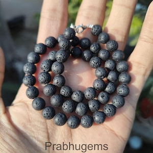 May include: A black lava stone bead necklace with a silver clasp. The necklace is made up of many round, porous beads. The text "Prabhugems" is visible at the bottom of the image. The necklace is held in a person's hand.