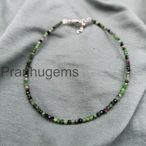 May include: An anklet featuring small, round beads in shades of green, black, and maroon. The beads are strung together with a silver-colored clasp. The anklet is displayed on a gray fabric surface. The text "Pranhugems" is visible.