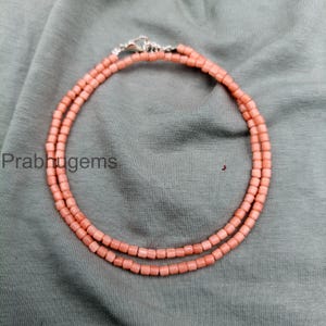 Könnte beinhalten: Eine Halskette aus kleinen, zylindrischen, pfirsichfarbenen Perlen, die in einem doppelten Kreis angeordnet sind. Die Kette hat einen silberfarbenen Metallverschluss oben und enthält den Text prabhugems auf der linken Seite.