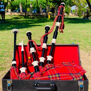 Puede incluir: Un juego de gaitas negras en un estuche negro con interior rojo. Las gaitas tienen cuerpos negros con detalles blancos y negros, cuerdas rojas y una bolsa de tartán roja y amarilla. El estuche está abierto.