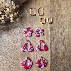 May include: An assortment of handmade earrings in various shapes and designs. The collection includes gold hoop earrings, flower-shaped earrings with red and white patterns, triangle earrings, and teardrop-shaped earrings. The earrings are displayed on a wooden surface.