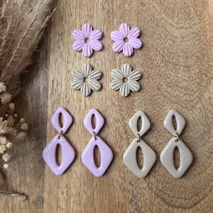 Può includere: Una collezione di quattro paia di orecchini. Due paia hanno la forma di fiori a sei petali in viola chiaro e beige. Due paia presentano un design geometrico a doppio rombo con un rombo superiore più piccolo e uno inferiore più grande.