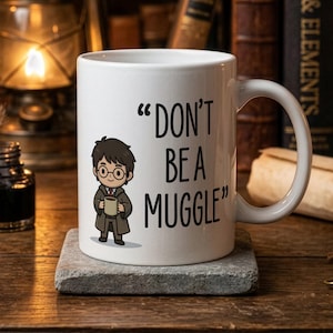 May include: White ceramic mug with the text "DON'T BE A MUGGLE" and a cartoon illustration of a character wearing glasses and holding a book. The mug sits on a gray stone and is surrounded by books and a lit candle.