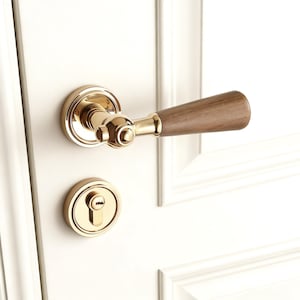 Peut inclure: Une poignée de porte et une serrure dorées avec une poignée en bois. La poignée a un design incurvé et est montée sur une porte blanche. Le trou de serrure est rond et également doré.