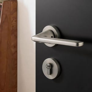 May include: A brushed nickel door handle and lockset are mounted on a black door. The handle has a curved design, and the lockset includes a keyhole. The door is positioned next to a white and brown wooden surface.