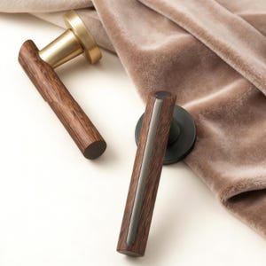 Könnte beinhalten: Zwei Türgriffe aus Holz und Metall. Ein Griff hat eine Messingoberfläche, der andere eine dunkle Metalloberfläche. Beide haben ein dunkles Holzmaserungsmuster. Die Griffe sind vor einem weichen, neutralfarbenen Stoff platziert.