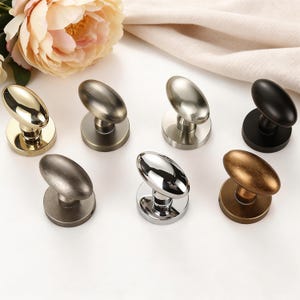 Peut inclure: Collection de sept boutons de meubles ovales dans différentes finitions métalliques, dont l'or, l'argent, le bronze et le noir. Chaque bouton a une base circulaire et une surface lisse et polie. Les boutons sont disposés sur une surface blanche.