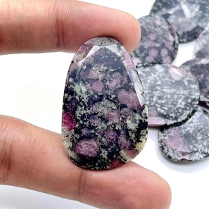 Peut inclure: Une pierre précieuse ovale et facettée avec un motif tacheté foncé de noir, de gris et de rose. La pierre polie est tenue entre les doigts, avec d'autres pierres similaires visibles en arrière-plan. La gemme a un aspect naturel et organique.