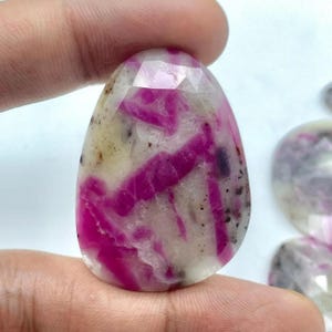 May include: A polished, oval gemstone with a faceted top. The stone displays a mix of white, beige, and vibrant pink hues, with streaks and inclusions creating a unique pattern. The gemstone is held between fingers.