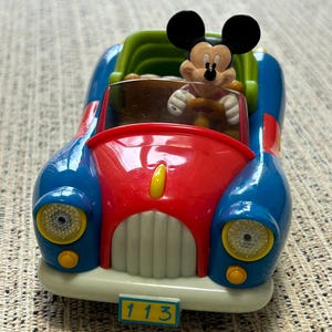 Op de afbeelding: Een kleurrijke speelgoedauto met Mickey Mouse achter het stuur. De auto heeft een blauwe carrosserie, een rode motorkap en een groene stoel. De nummerplaat toont "113". De koplampen zijn geel en de bumper is crèmekleurig.