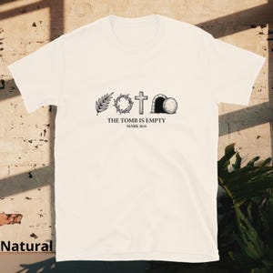 May include: A cream-coloured t-shirt with a graphic depicting a palm branch, a crown of thorns, a cross, and an empty tomb. The text "THE TOMB IS EMPTY" and "MARK 16:6" are printed below the images. The shirt is made of a soft material.