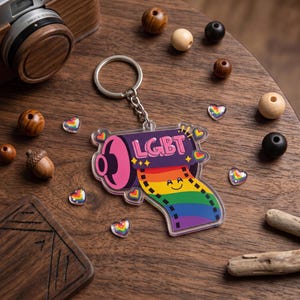 May include: A clear acrylic LGBT keychain with a rainbow film strip design. The keychain has a silver chain and ring. The word "LGBT" is written in pink and yellow. The keychain is surrounded by heart-shaped gems.
