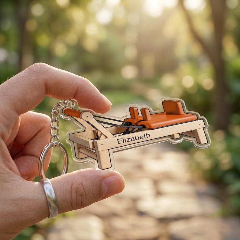 May include: A keychain featuring a miniature Pilates reformer machine. The keychain is made of clear acrylic with a wooden frame, orange accents, and the name "Elizabeth" printed on it. The keychain is attached to a silver chain and ring.