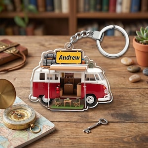 May include: A red and white camper van keychain with the name "Andrew" on a yellow sign. The keychain has a silver key ring and chain. The van has an open side with miniature furniture inside. A compass and key are also visible.