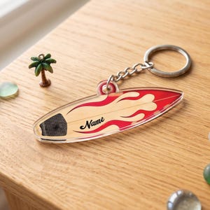 May include: A surfboard-shaped keychain with a red flame design and the word "Name" in black script. The keychain is attached to a silver chain and ring. A small palm tree figurine is next to the keychain on a wooden surface.