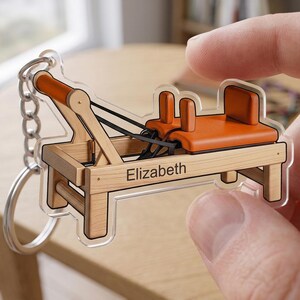 May include: A clear acrylic keychain featuring a miniature Pilates reformer machine. The keychain has a wooden frame with orange accents and the name "Elizabeth" printed on it. The keychain is attached to a silver chain.