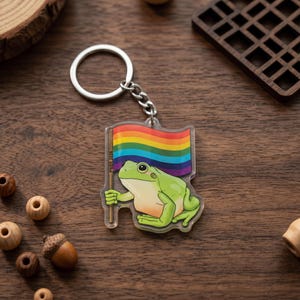 May include: A clear acrylic keychain featuring a green frog holding a rainbow flag. The keychain has a silver metal ring and chain. The frog has a white belly and is holding a flag with the text "PRIDE 2024".