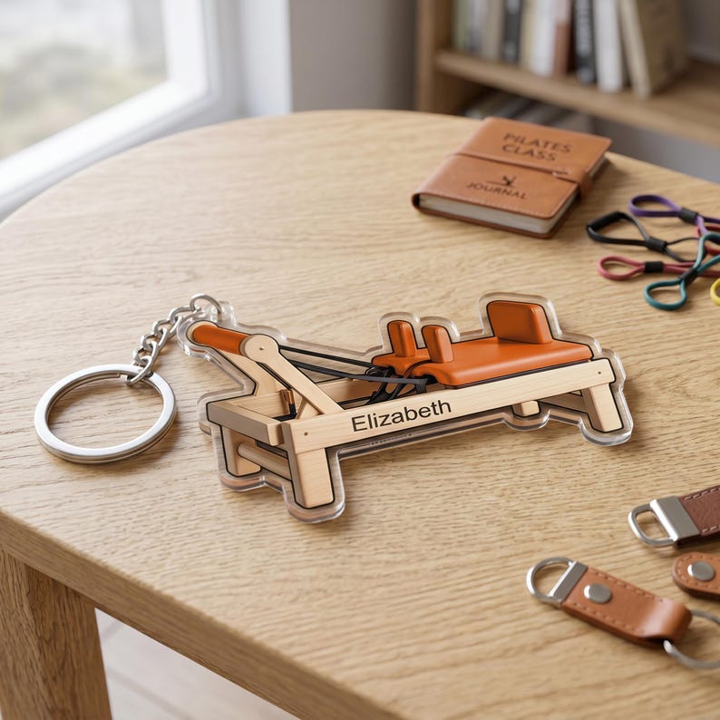 May include: A clear acrylic keychain shaped like a Pilates reformer machine, with a silver keyring. The machine has a light brown wooden frame, orange seat and details, and the name "Elizabeth" is printed on it.