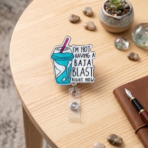 May include: A badge reel with a Baja Blast design. The badge features a turquoise and white drink with a pink straw and the text "I'M NOT HAVING A BAJA BLAST RIGHT NOW". The reel has a clear plastic strap and a silver button.