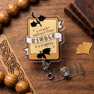 May include: A clear acrylic badge reel with a rectangular design. The badge has a tan background with black dragon silhouettes and the text "a reader without their KINDLE is a tragedy". The reel has a clear plastic strap and a metal clip.
