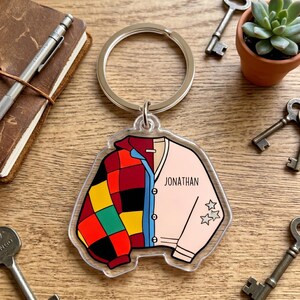 May include: A clear acrylic keychain featuring a graphic of a split-design jacket. The left side has a colorful red, yellow, green, and black checkered pattern, while the right side is light pink with the name JONATHAN and three small star icons.