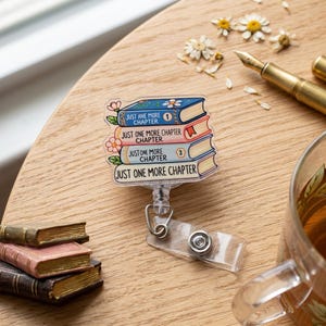 May include: A clear badge reel with a stack of books design. Each book is a different color and has the text "JUST ONE MORE CHAPTER". The badge reel is on a wooden surface with a pen and flowers.