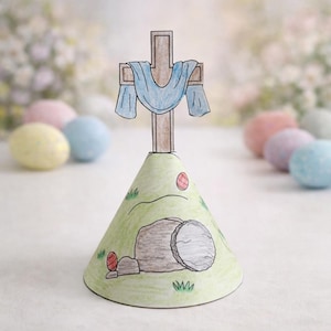 May include: A handmade Easter-themed paper craft featuring a brown cross with a blue cloth draped over it, atop a green hill with a tomb and Easter eggs. The craft is designed with a cone shape.