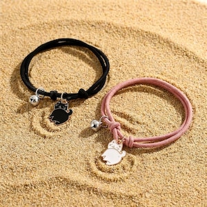 May include: Two matching cat charm bracelets. One bracelet is black with a black cat charm, and the other is pink with a white cat charm. Each bracelet has a small silver bell. The bracelets are on a sandy surface.