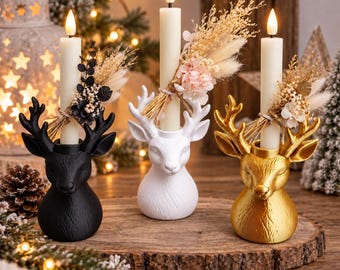 Puede incluir: Tres portavelas con forma de cabeza de ciervo en negro, blanco y dorado, cada uno con una vela cónica larga de color crema. Cada vela está adornada con un pequeño ramo de flores secas y hierbas atado con cordel, creando un conjunto decorativo.
