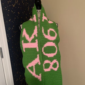May include: A green crocheted scarf with pink lettering that reads "Hufflepuff".