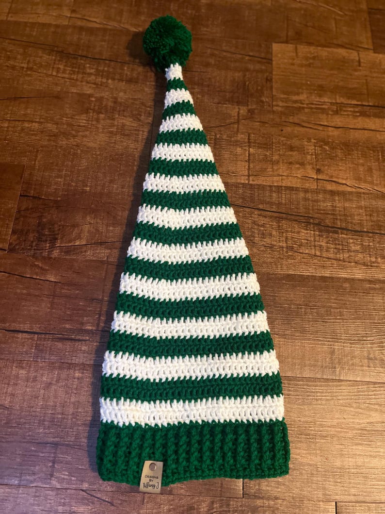 May include: Green and white striped crochet elf hat with a white pom pom on top. The hat has a tag that says "Designed by Tiffany J".