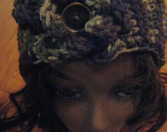 May include: A crocheted headband in a dark blue, green and white colour scheme. The headband features a large, round, brown button in the centre.