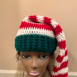 May include: A red, white and green crocheted elf hat with a long, striped tail. The hat is made with a textured stitch and has a pointed top.