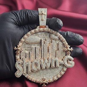 May include: A custom pendant with the name "Shawn-G" in a cityscape design. The pendant is encrusted with numerous small, sparkling stones, set in a gold-colored metal. A cross-shaped element is at the top.