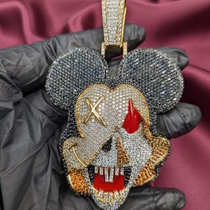 May include: A pendant featuring a skull design with a Mickey Mouse silhouette. The pendant is encrusted with black, white, and gold-colored gemstones. The skull has a red eye and a gold cross over one eye. The pendant is held by a gold bail.