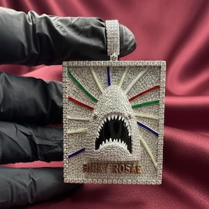 Peut inclure: Pendentif incrusté de diamants représentant un requin avec une gueule ouverte et des dents acérées. Le pendentif rectangulaire présente des rayons colorés émanant de la tête du requin. Le texte "RICKY ROSAE" est en bas.