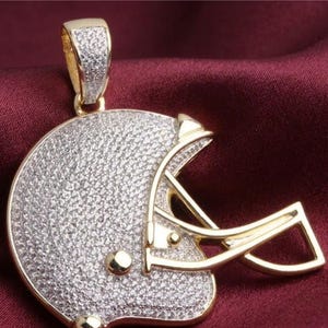 May include: A gold-toned pendant in the shape of a football helmet, encrusted with small, sparkling stones. The helmet features a gold-colored face mask and a bail for a chain. The pendant is set against a burgundy background.