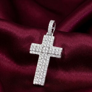 Peut inclure: Un pendentif en forme de croix argentée, incrusté de nombreux petits pierres précieuses transparentes. La croix est suspendue à une bélière assortie. Le pendentif est posé sur un fond texturé rouge foncé.