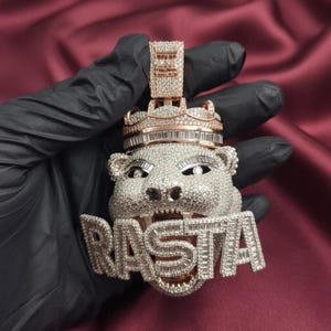 May include: A diamond-encrusted pendant featuring a roaring bear design with a crown. The word "RASTA" is spelled out in large, sparkling letters below the bear's face. The pendant has a gold and silver color scheme.