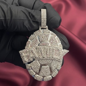May include: A silver pendant with a geometric design, encrusted with small, sparkling stones. The pendant has a bale for a chain and features the letters "AM" in a stylized font. The overall shape is a rounded, shield-like form.