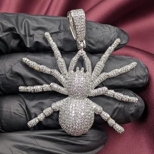 May include: A silver-colored pendant in the shape of a spider, encrusted with small, sparkling stones. The spider has eight articulated legs and a detailed body. The pendant is held by a silver bail. The background is a dark, textured surface.
