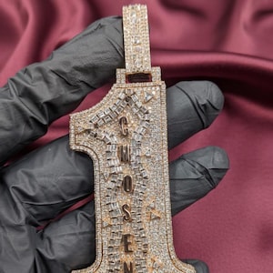 May include: A gold-toned pendant in the shape of the number one, encrusted with numerous small, sparkling stones. The word "CHOSEN" is spelled out vertically in the center of the pendant. The pendant is held by a gloved hand.