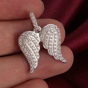 Peut inclure: Un pendentif argenté en forme d'ailes d'ange, incrusté de nombreuses petites pierres scintillantes. Les ailes sont détaillées avec des lignes de plumes individuelles. Une petite boucle en haut permet de l'attacher.