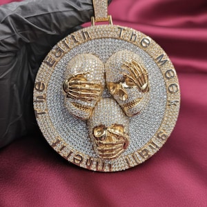 Op de afbeelding: Een goudkleurige hanger met drie schedelfiguren, elk met een hand die de ogen, mond en oren bedekt. De hanger is bezet met kleine, sprankelende stenen en heeft de tekst "The Earth in The Meek" langs de rand.