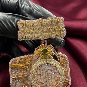 May include: A gold-toned pendant featuring a camera design, encrusted with numerous small, clear gemstones. The pendant has a rectangular top piece with the text "Show Me The Money" also covered in gemstones. The background is a deep red.