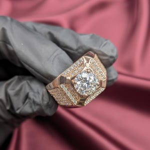 Może przedstawiać: Pierścionek z różowego złota z dużym, przezroczystym kamieniem centralnym o okrągłym szlifie. Pierścionek jest inkrustowany mniejszymi, przezroczystymi kamieniami wokół środka i na obrączce. Pierścionek jest trzymany na różowym tle.