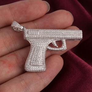 May include: A silver-colored pendant in the shape of a handgun, encrusted with numerous small, clear gemstones. The pendant is held in a person's hand against a burgundy background. The handgun pendant is a jewelry item.