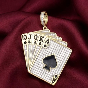 May include: A gold-toned pendant featuring a hand of playing cards: 10, Jack, Queen, King, and Ace of Spades. The cards are encrusted with small, clear stones, and the spade symbol is black. The pendant has a gold bail.
