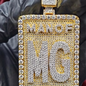 May include: Gold-toned pendant with the words "MAN OF GOD" and the initials "MG" encrusted with sparkling white stones. The pendant has a rectangular shape with a rectangular bail, and is set against a dark background.
