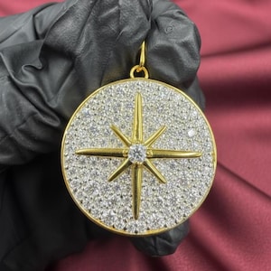 May include: A gold-colored pendant with a starburst design. The pendant features a central, clear gemstone surrounded by a gold star design. The background is covered in small, clear gemstones. The pendant is round and has a gold bail.